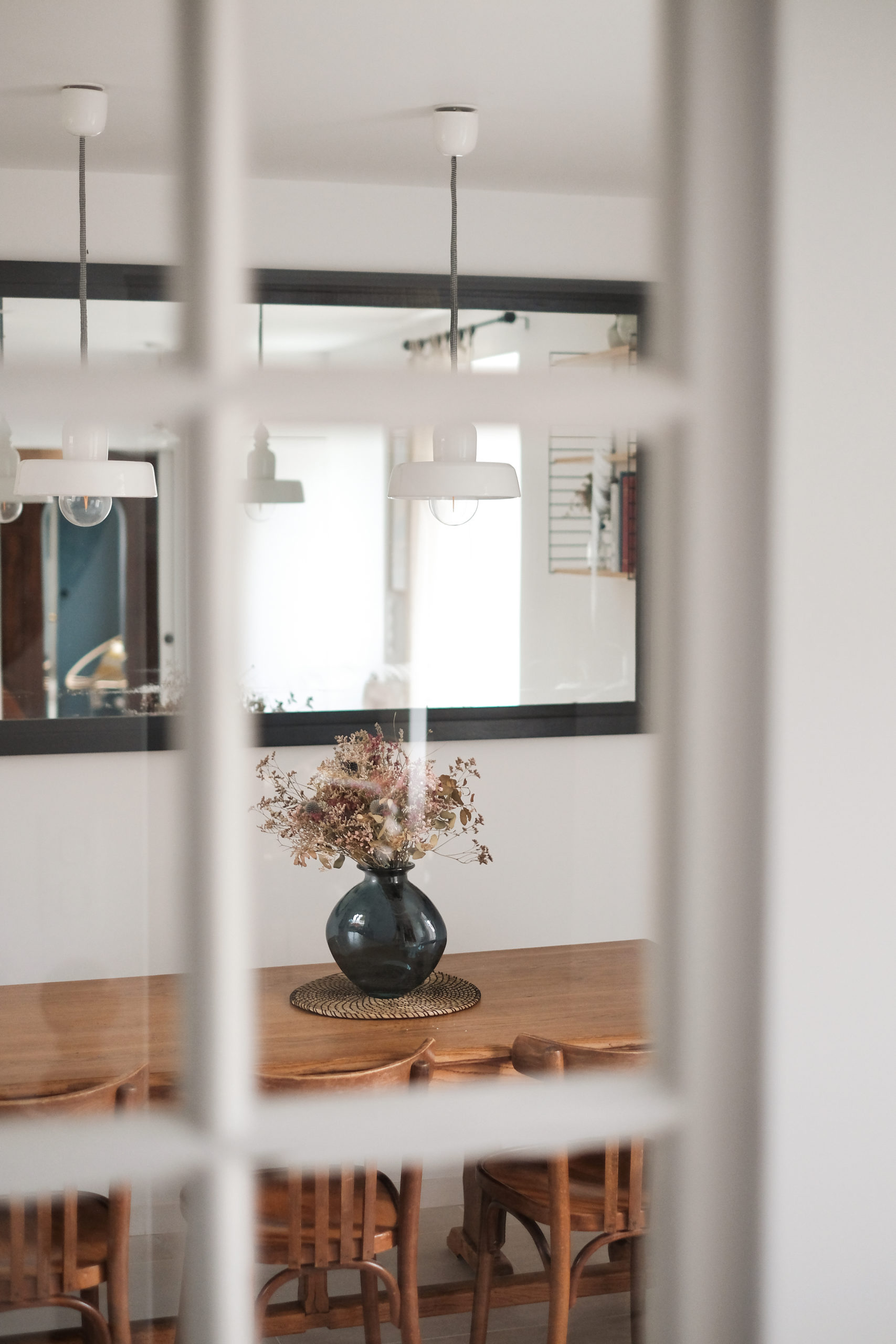 On aperçoit une table en bois surmontée d'un vase bleu à travers une fenêtre